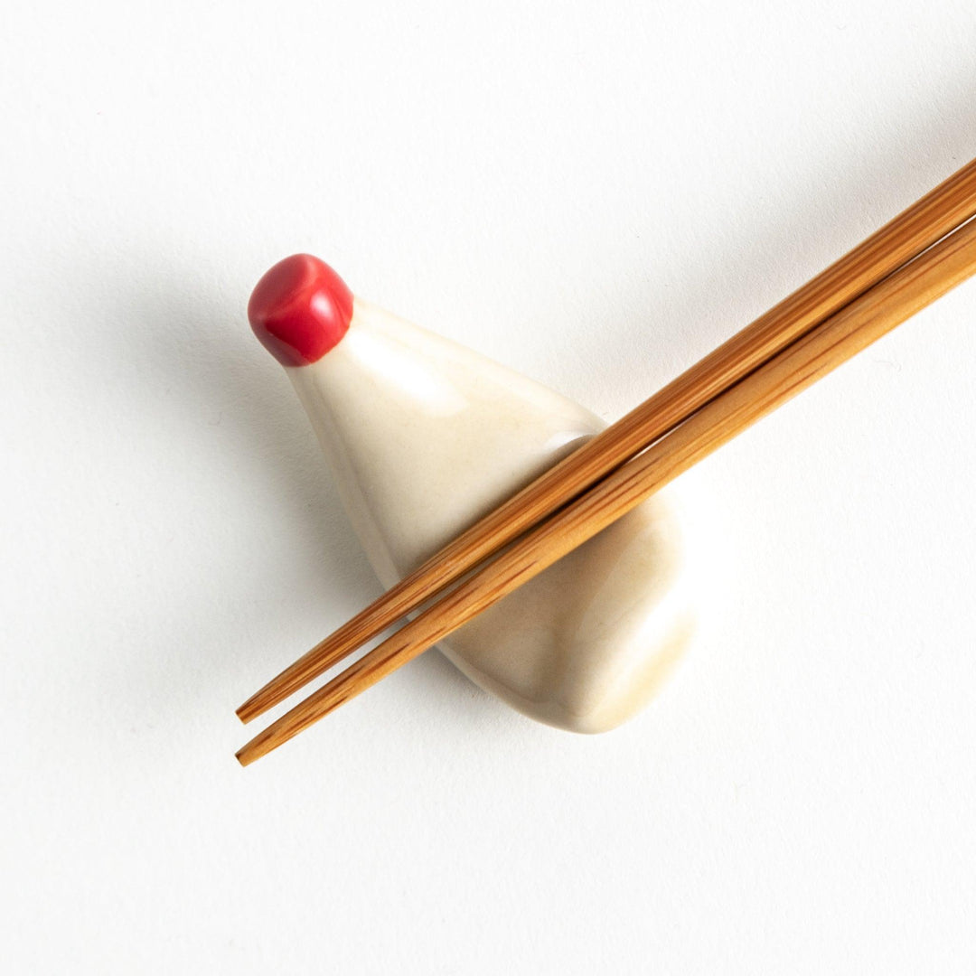 A small ceramic chopstick rest shaped like a mayonnaise bottle with a red cap, holding a pair of wooden chopsticks, placed on a white background.