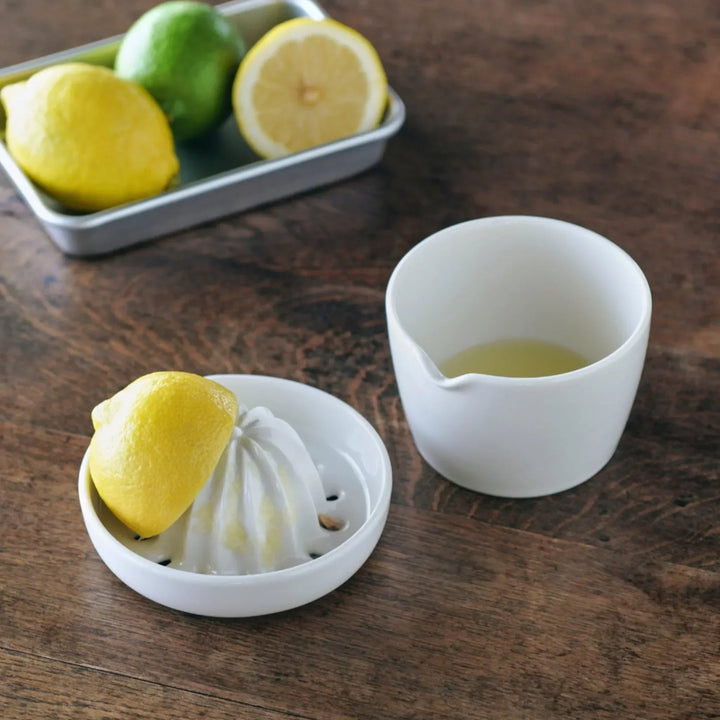 White ceramic citrus juicer with a half lemon on top, paired with a small spouted cup containing freshly squeezed juice.
