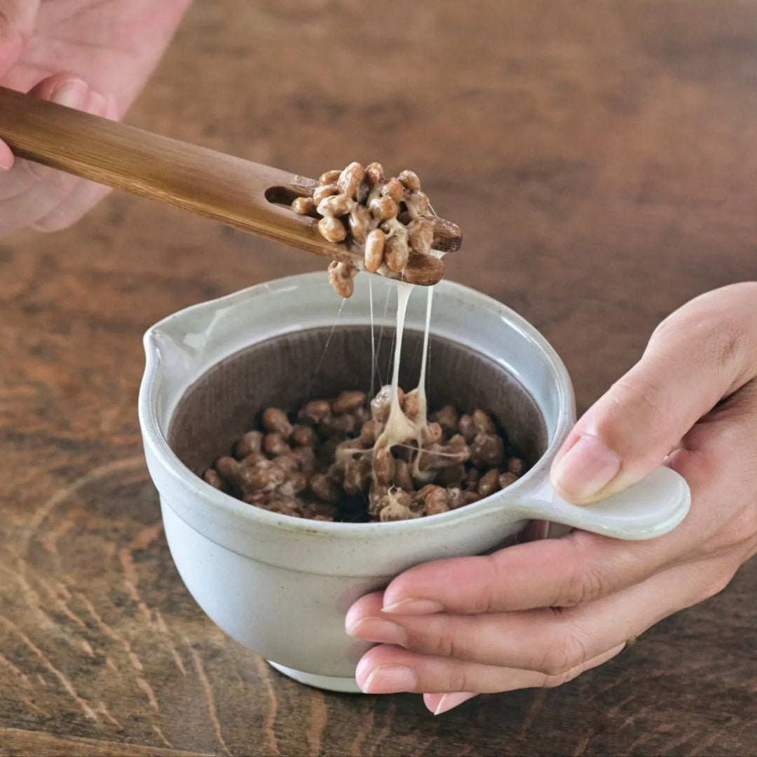 White ceramic suribachi bowl with spout, paired with a bamboo stirring stick lifting sticky natto beans.