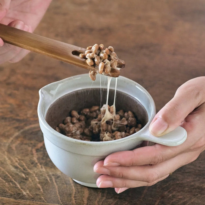 White ceramic suribachi bowl with spout, paired with a bamboo stirring stick lifting sticky natto beans.