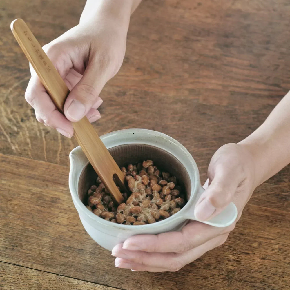 Hand stirring natto beans in a white ceramic suribachi bowl with a bamboo stick, placed on a wooden table.