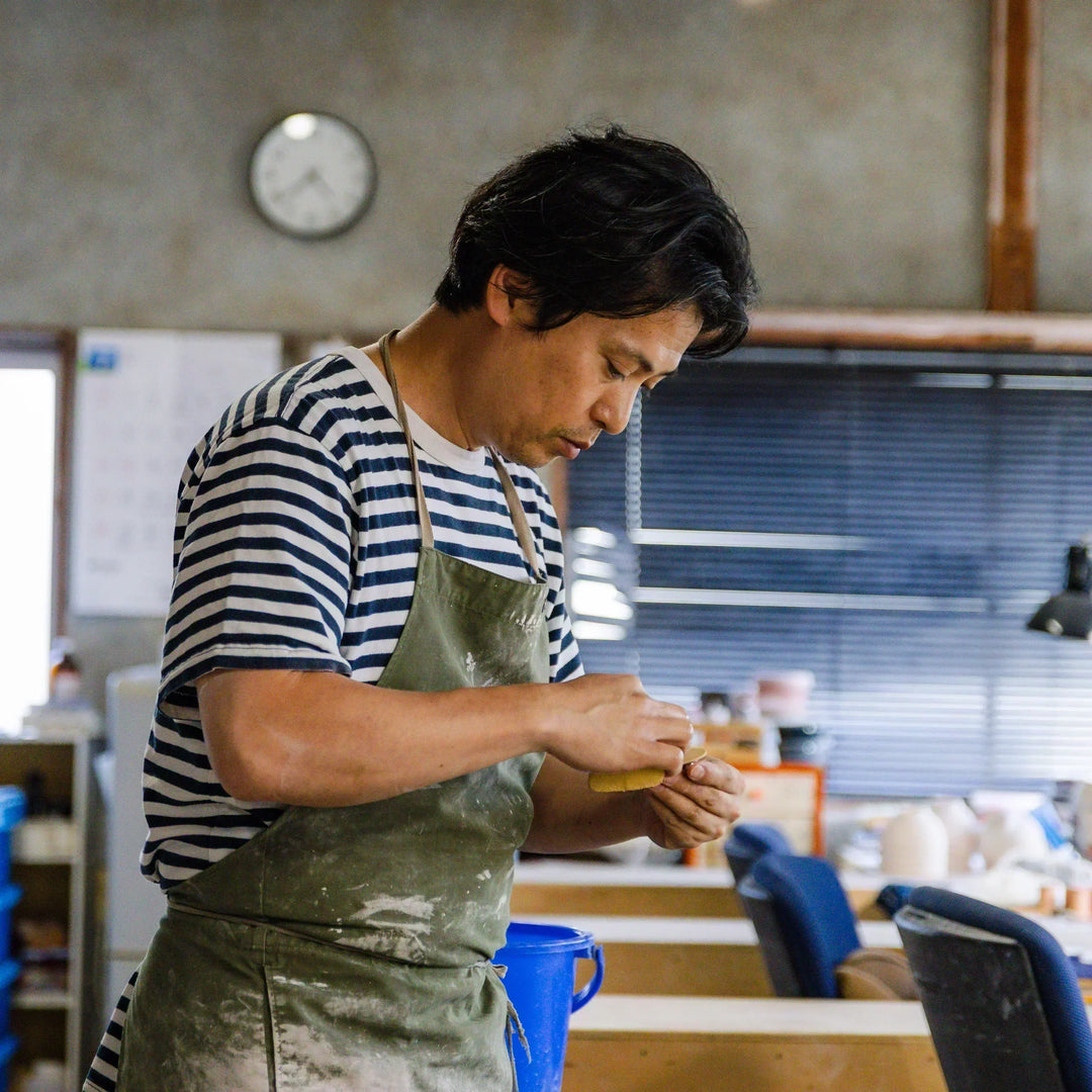 A ceramic artisan shaping clay by hand at a workbench, wearing a striped shirt and apron inside a calm, tool-filled workshop