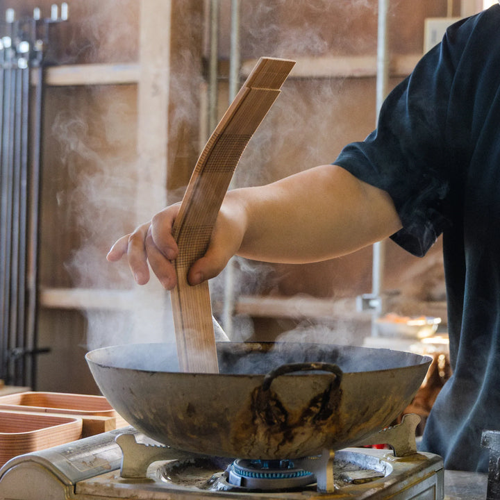 Steam rises as a craftsperson heats and bends a carved wooden board over a pot, showing a step in traditional woodworking