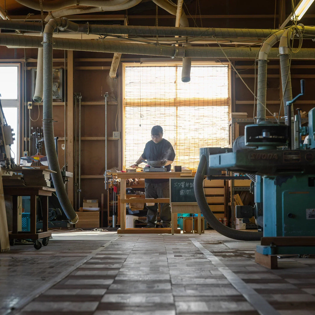 A craftsperson working at a bench in a spacious woodworking studio filled with tools machines and natural light