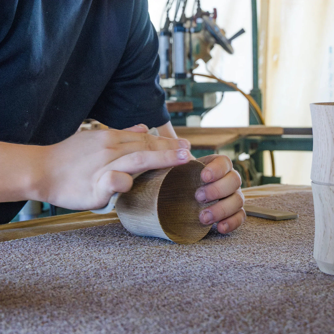 Hands sanding a wooden cup in a workshop, refining the surface with tools in the background