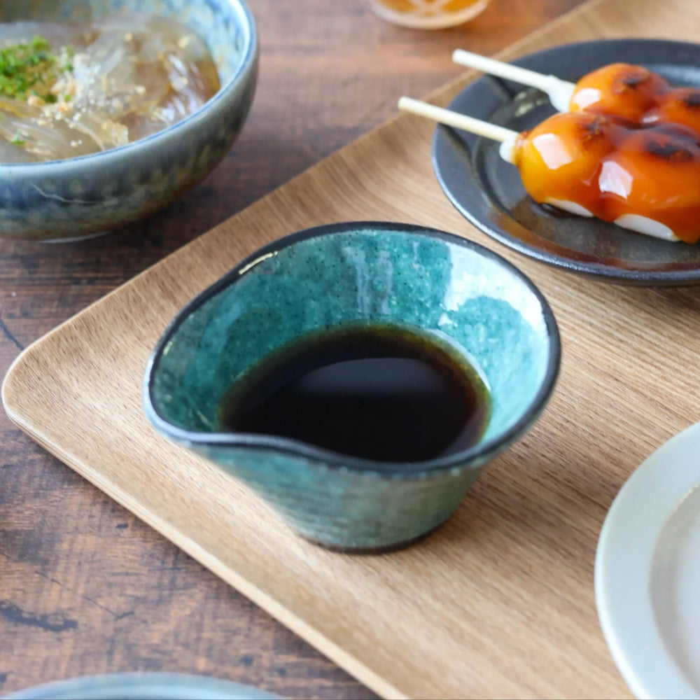 Wavy edged blue green condiment bowl with soy sauce on wooden tray beside sweets and chilled noodles.