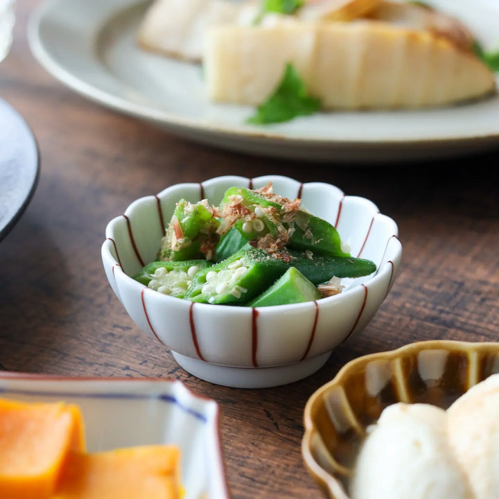 A chrysanthemum-shaped porcelain Japanese condiment bowl with vertical red lines, serving sliced okra.