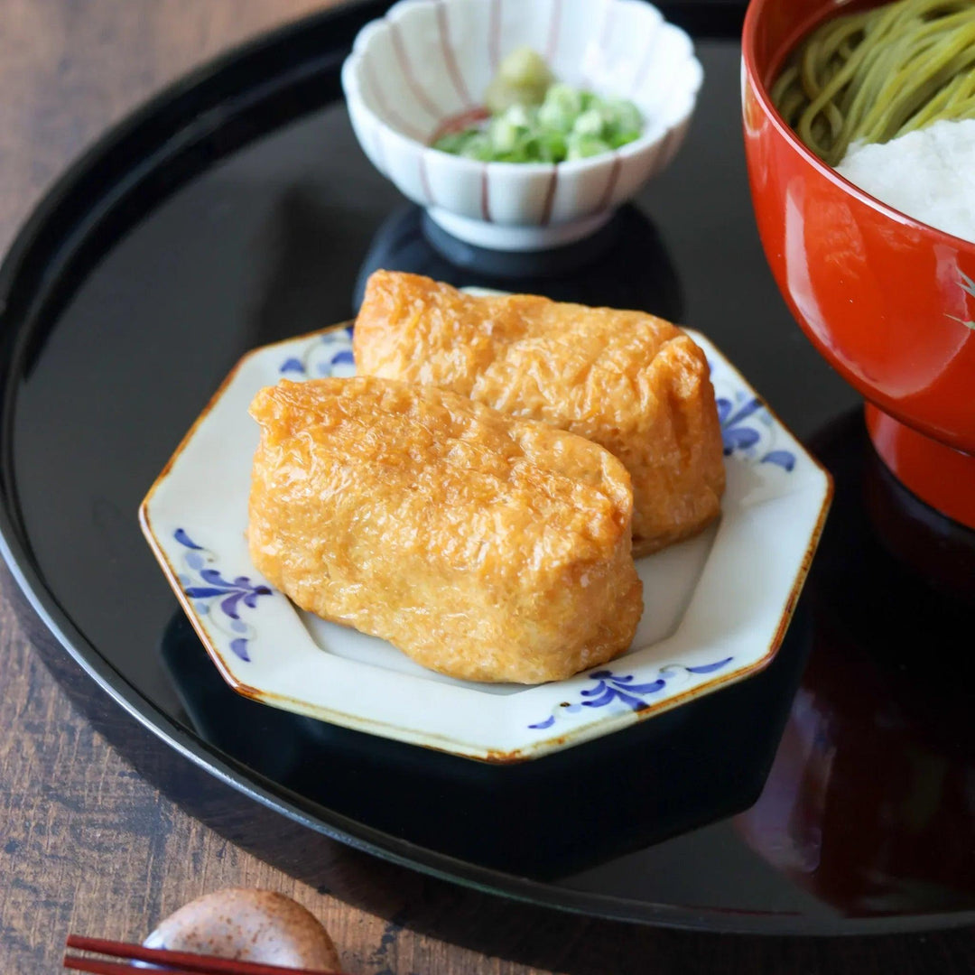 Two inari sushi served on an octagonal porcelain Japanese soy sauce dish with blue floral leaf patterns and gold trim.