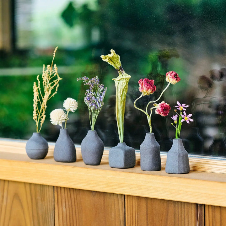 Black minimalist ceramic bud vases in geometric shapes on a wooden windowsill holding dried wildflowers and pods as decor.