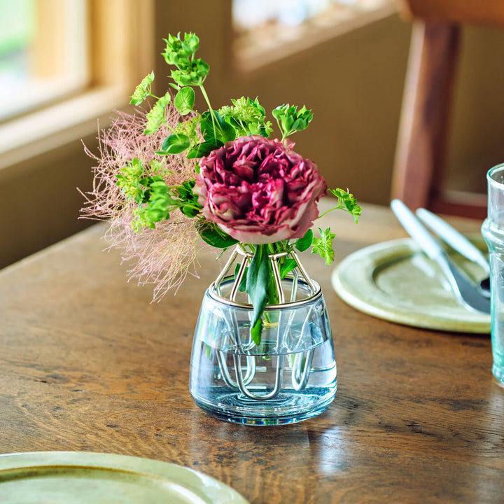 Glass bud holder with brass grid insert showcasing deep pink ruffled rose and airy green sprigs in water on wooden table.