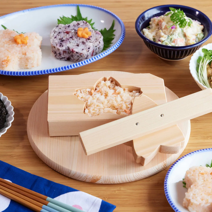 Sakura-shaped onigiri mold made of hinoki wood, displayed with rice balls, potato salad, and other side dishes.