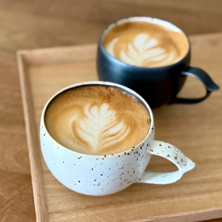 Speckled white and matte black ceramic mugs with latte art on wooden tray, highlighting artisanal Japanese tableware