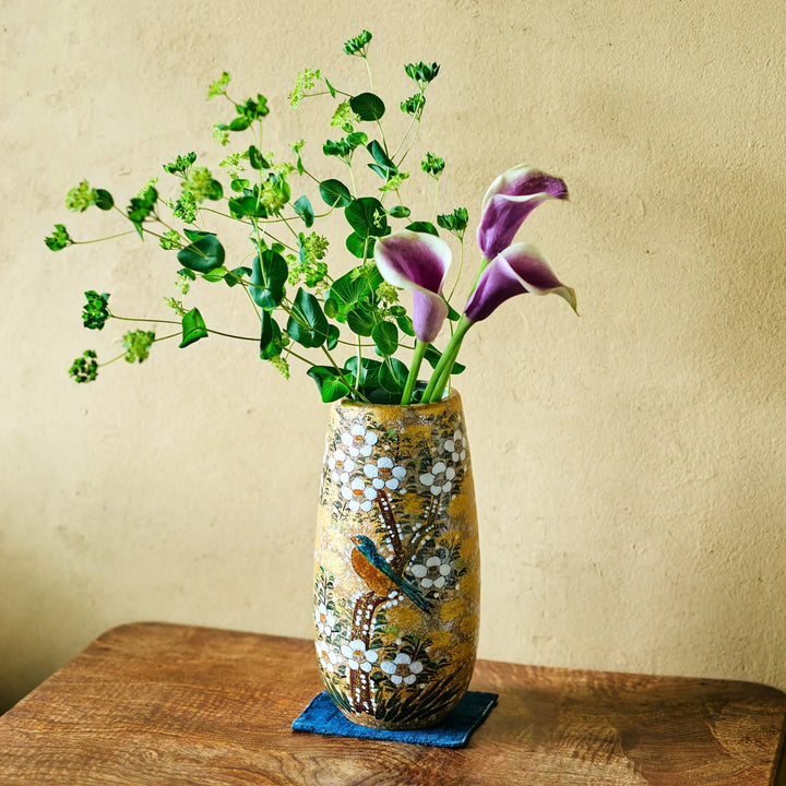 Gold-speckled ceramic vase with white blossoms and bird motif holding purple calla lilies and green sprigs on wooden tabletop