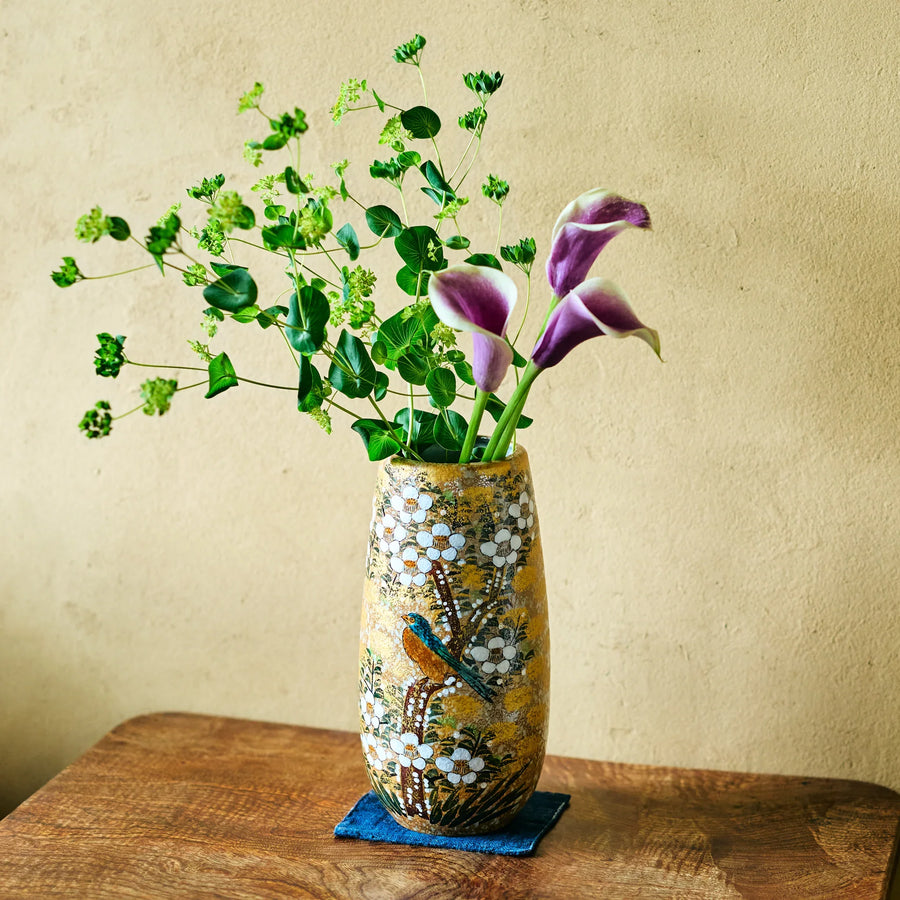 Gold-speckled ceramic vase with white blossoms and bird motif holding purple calla lilies and green sprigs on wooden tabletop