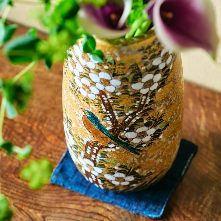 Close-up of gold-speckled vase featuring painted white blossoms and turquoise bird perched against textured background.
