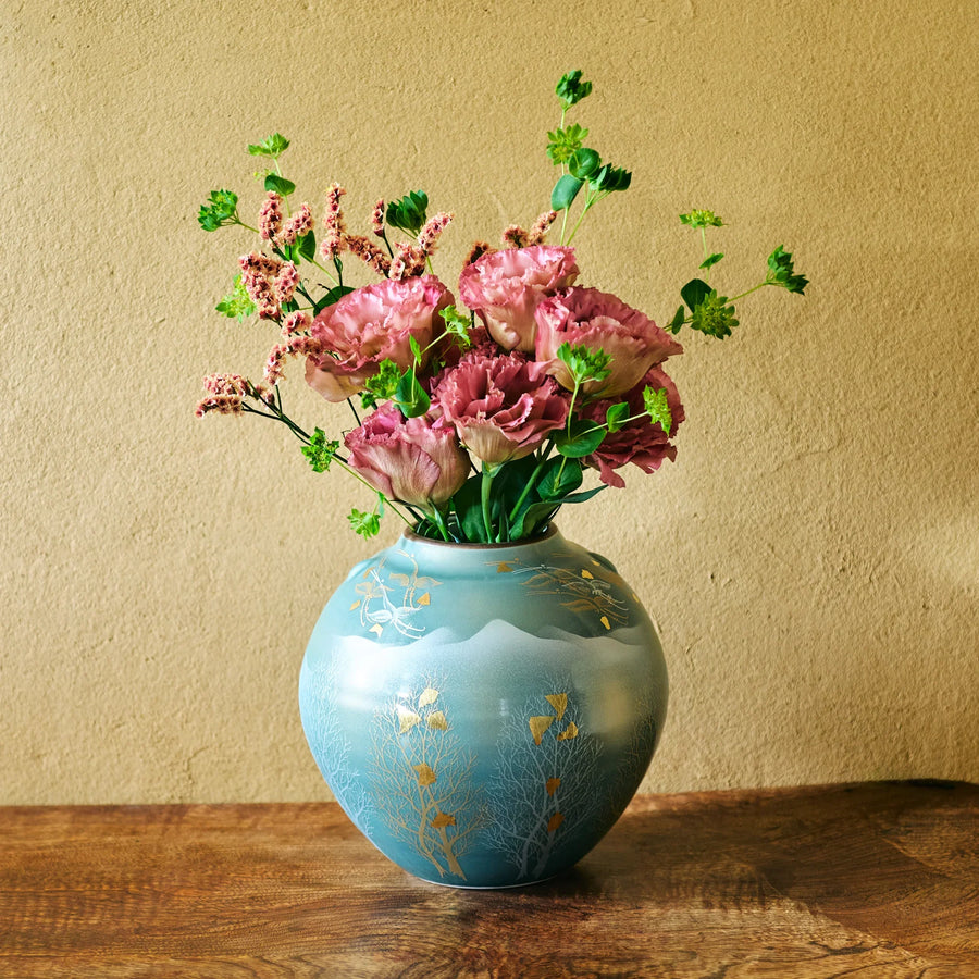 Hand-painted turquoise round vase with gold leaf motifs showcasing a lush bouquet of ruffled pink blooms and green foliage.