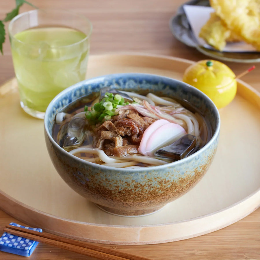 A rustic indigo ceramic donburi bowl filled with udon, garnished with green onions, kamaboko, and vegetables.