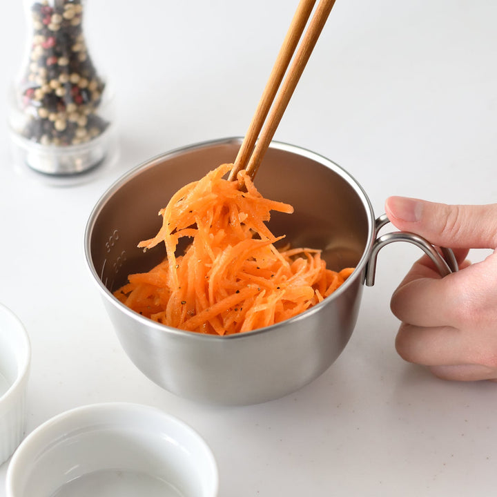 Person using chopsticks to lift shredded carrot salad out of a stainless steel egg beating bowl with handle