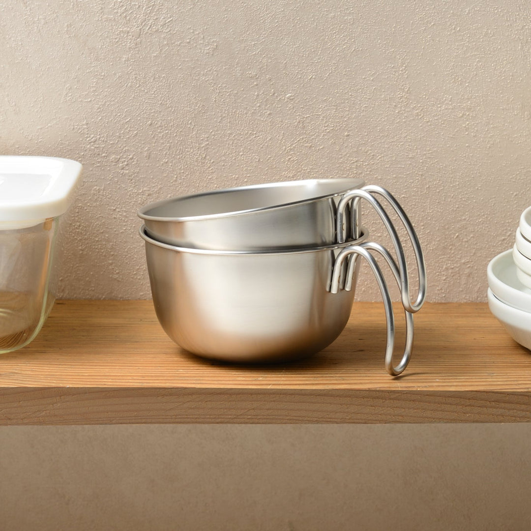 Two stainless steel egg beating bowls with handles neatly stacked for storage on a wooden shelf