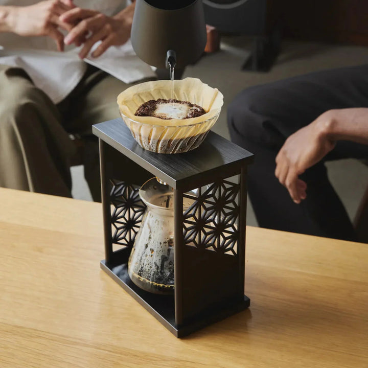 Pouring hot water from black kettle into hario aya asanoha black wood coffee dripper set with men relaxing in room