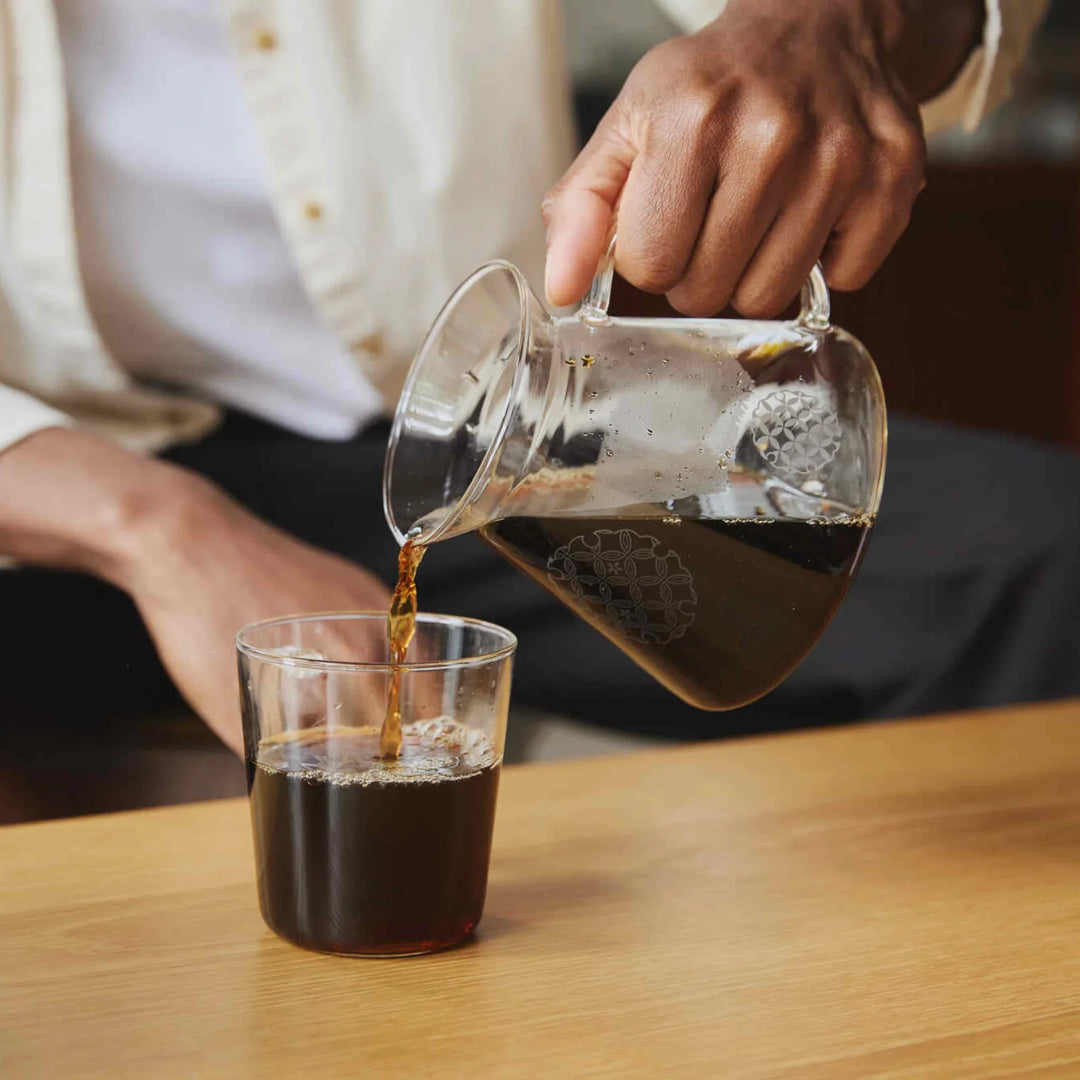 Pouring fresh coffee from patterned hario glass range server into clear drinking cup on wooden table coffeeware