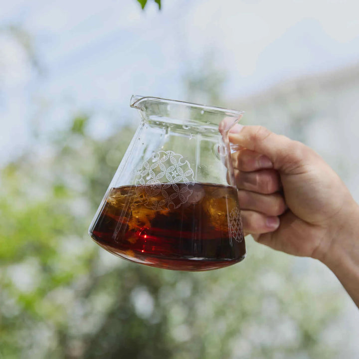 Holding hario patterned glass range server filled with iced coffee against bright outdoor garden background bokeh