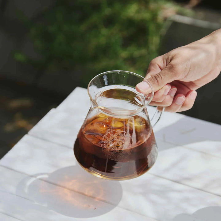Holding hario patterned glass range server with iced coffee casting colorful shadow on white wooden table outdoors