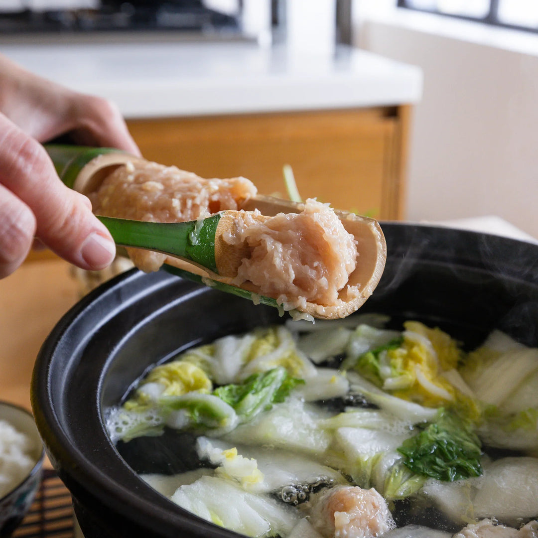 Hands using the bamboo tsumire server set to scoop freshly made chicken meatballs into a bubbling hot pot with vegetables