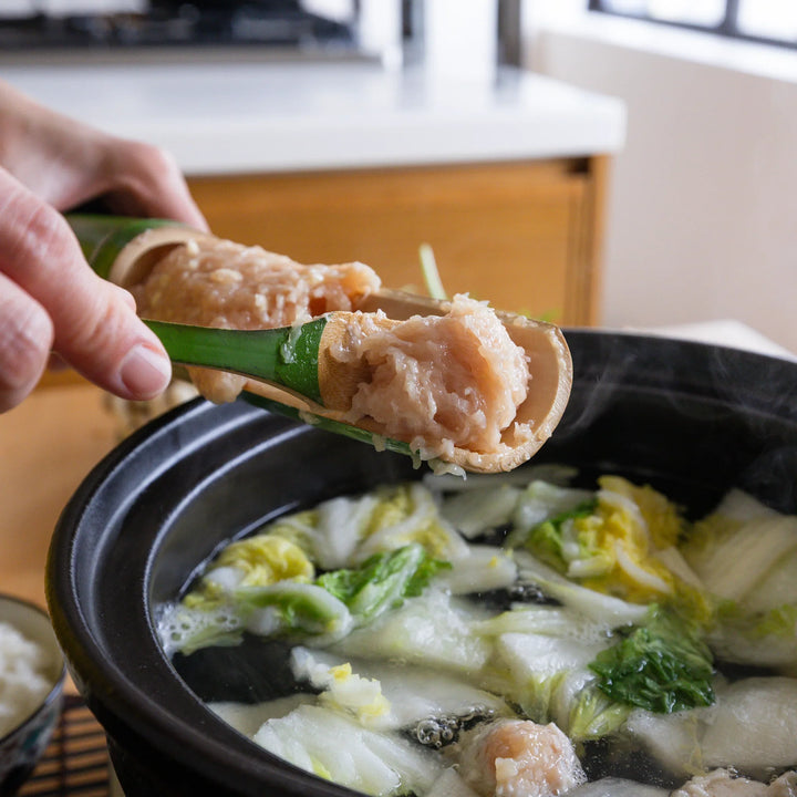 Hands using the bamboo tsumire server set to scoop freshly made chicken meatballs into a bubbling hot pot with vegetables