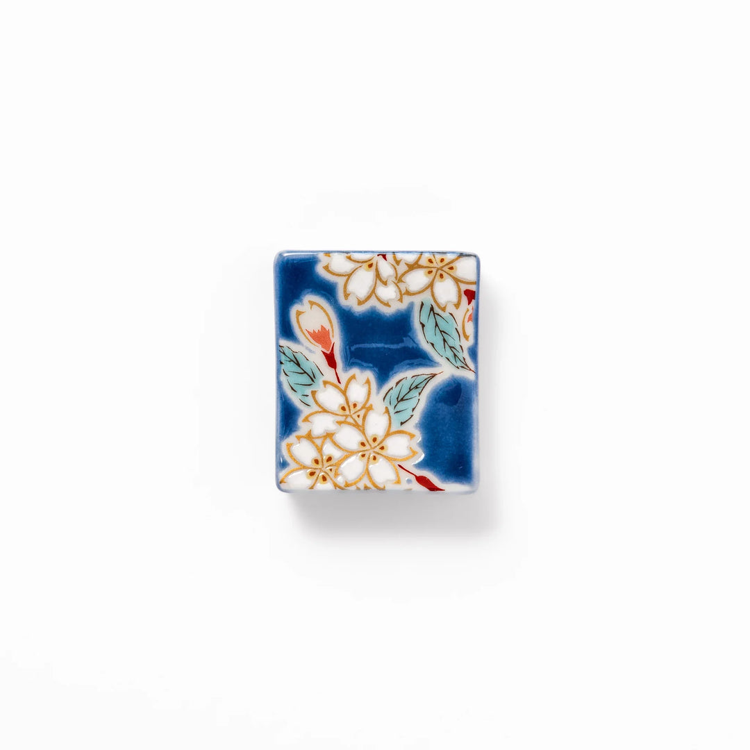 Rectangular blue Japanese ceramic chopstick rest with white sakura blossoms and green leaves for dining.