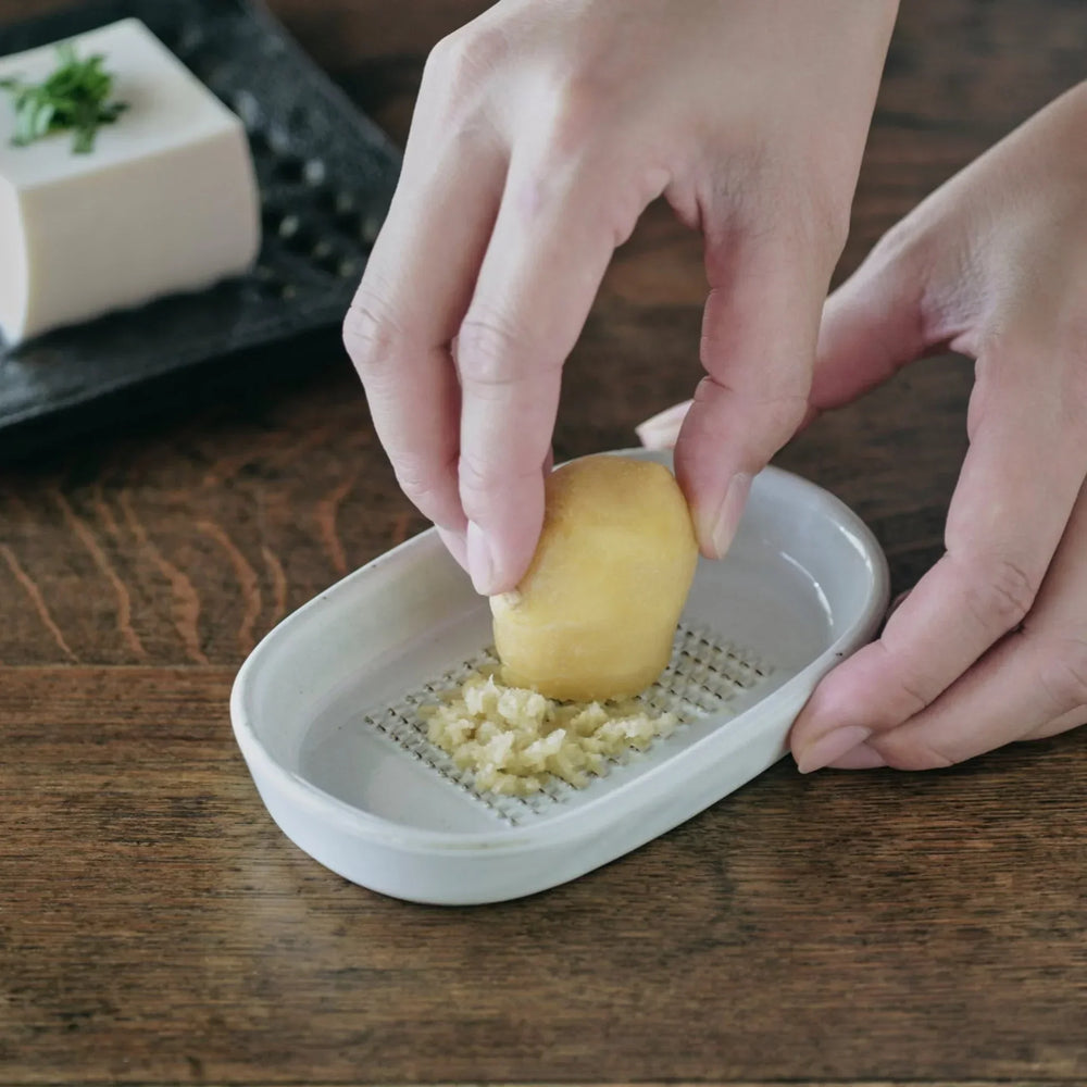 White ceramic ginger grater dish with raised teeth, shown in use grating fresh ginger on a wooden table.