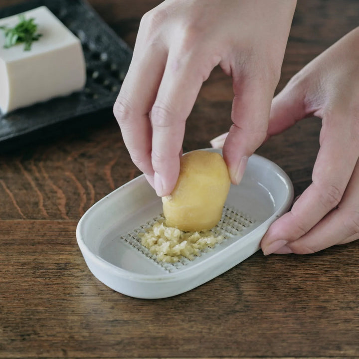 White ceramic ginger grater dish with raised teeth, shown in use grating fresh ginger on a wooden table.