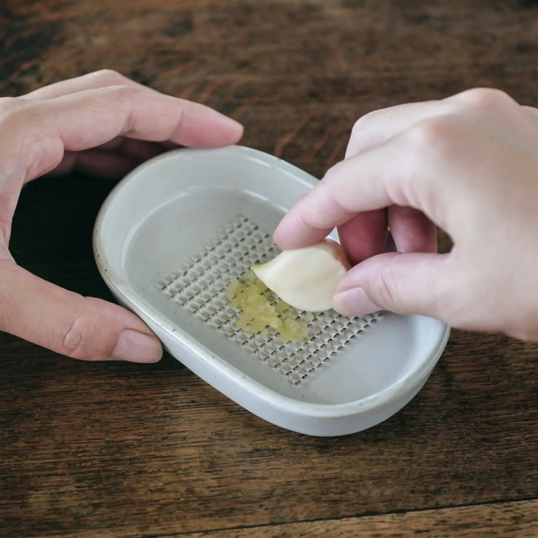 White ceramic garlic grater dish with raised teeth, shown in use grating fresh garlic on a wooden table.