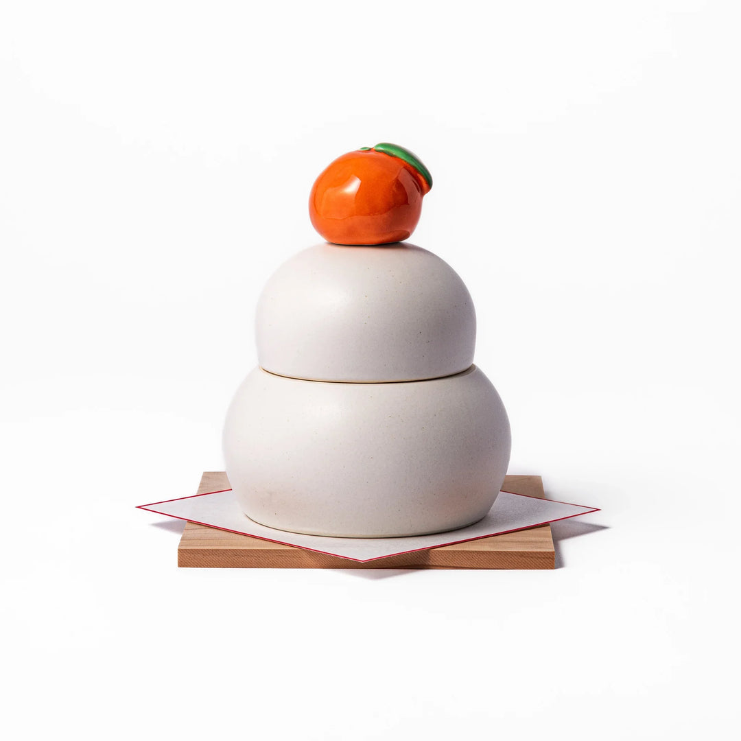 Ceramic kagami mochi jar with orange ornament on top, white two-tier design, and wooden tray for New Year decoration