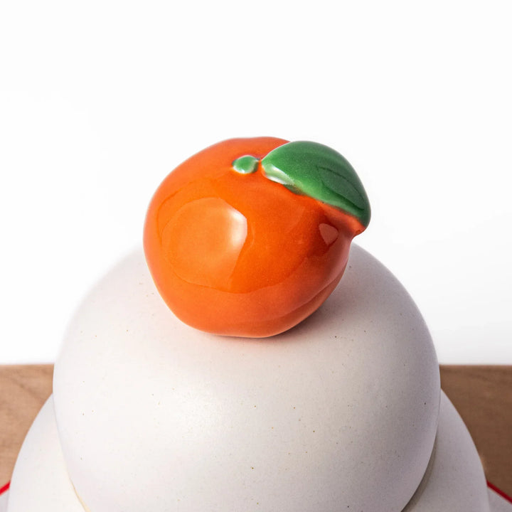 Ceramic kagami mochi jar with orange ornament on top, white two-tier design, and wooden tray for New Year decoration
