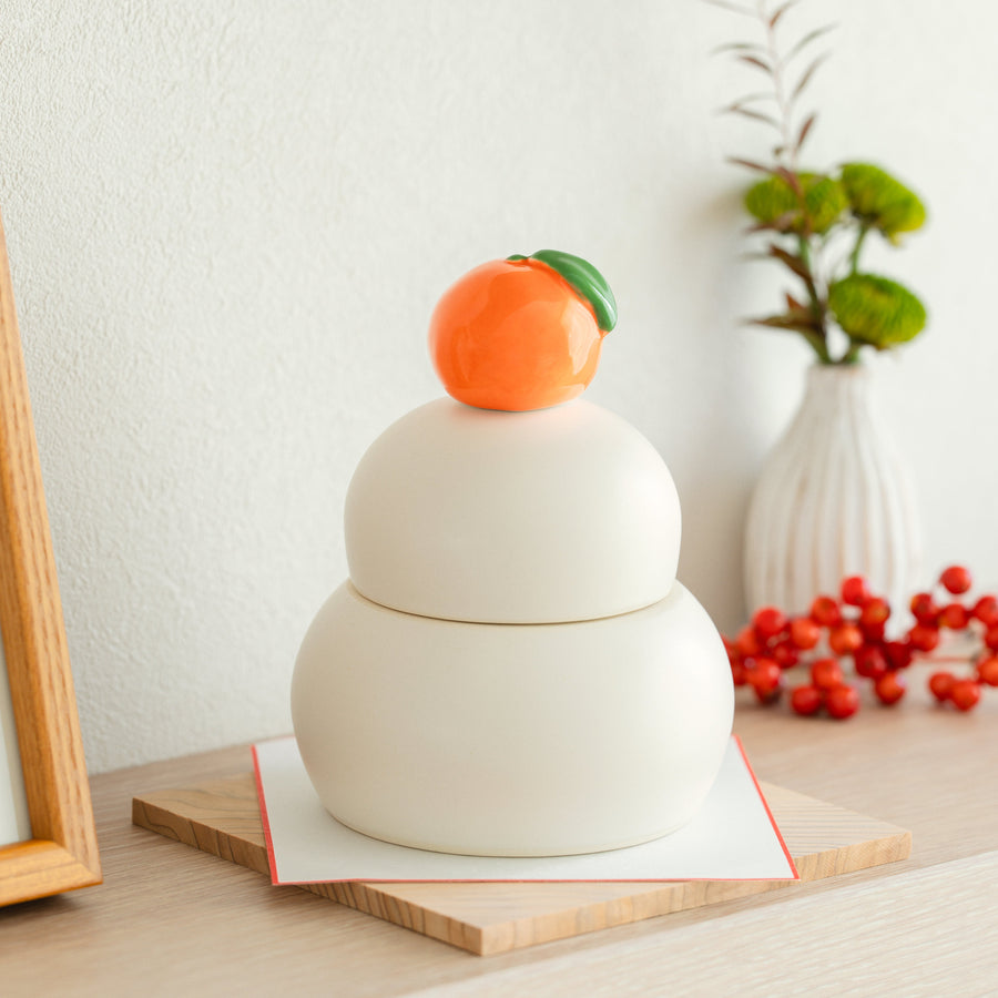 Side view of a white ceramic Japanese Kagami Mochi jar with an orange mandarin lid on a decorative wooden base