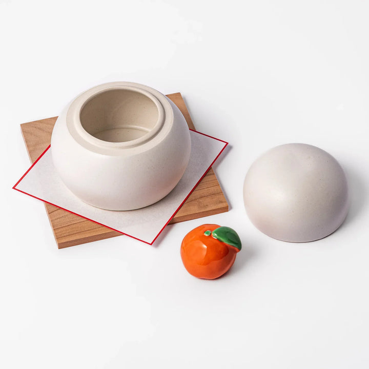 Ceramic kagami mochi jar with orange ornament on top, white two-tier design, and wooden tray for New Year decoration