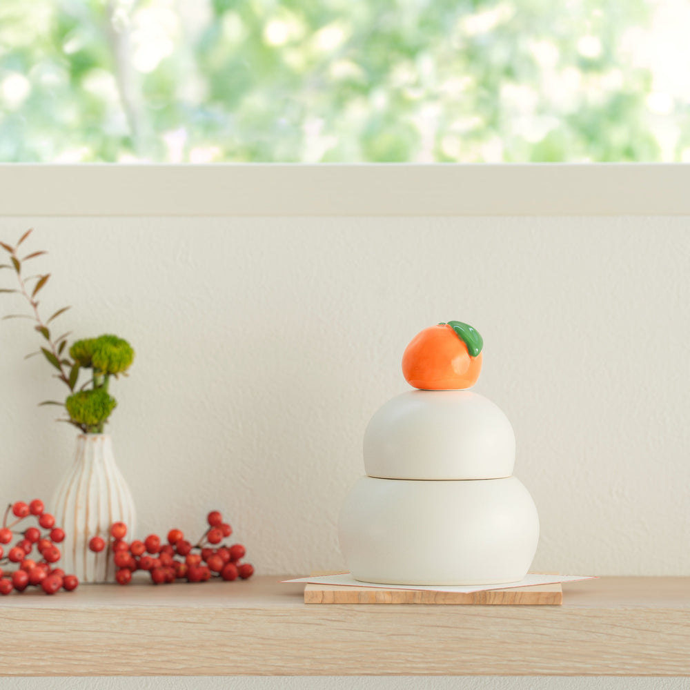 A white ceramic Japanese Kagami Mochi keepsake jar topped with a small orange mandarin on a wooden stand