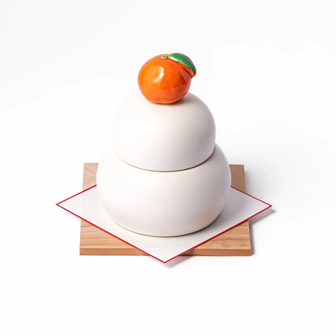 Ceramic kagami mochi jar with orange ornament on top, white two-tier design, and wooden tray for New Year decoration