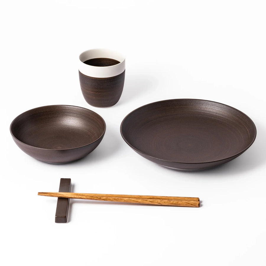 Ceramic bowl, plate, and cup set in rich brown with natural wood chopsticks and matching chopstick rest on a white background