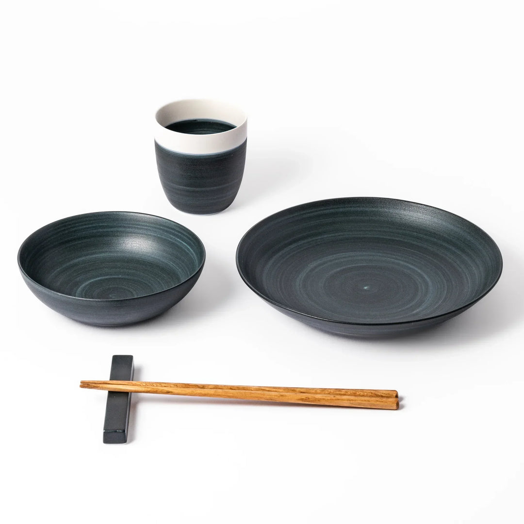 Japandi-style table setting in deep navy tones featuring a matching salad plate, bowl, tea cup, and chopsticks with rest.