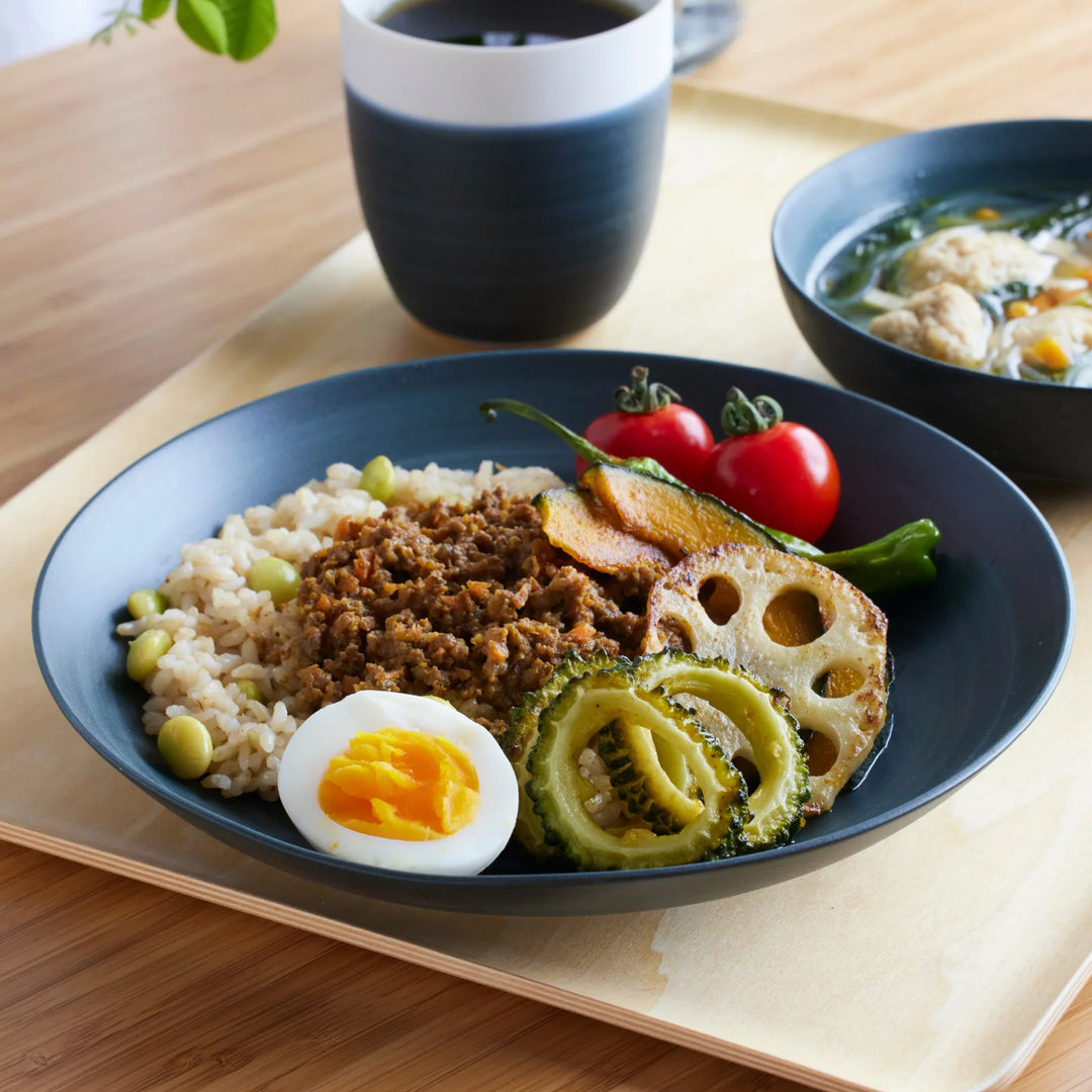 Navy blue salad plate with assorted vegetables, rice, and boiled egg, paired with matching soup bowl and cup on wooden tray.