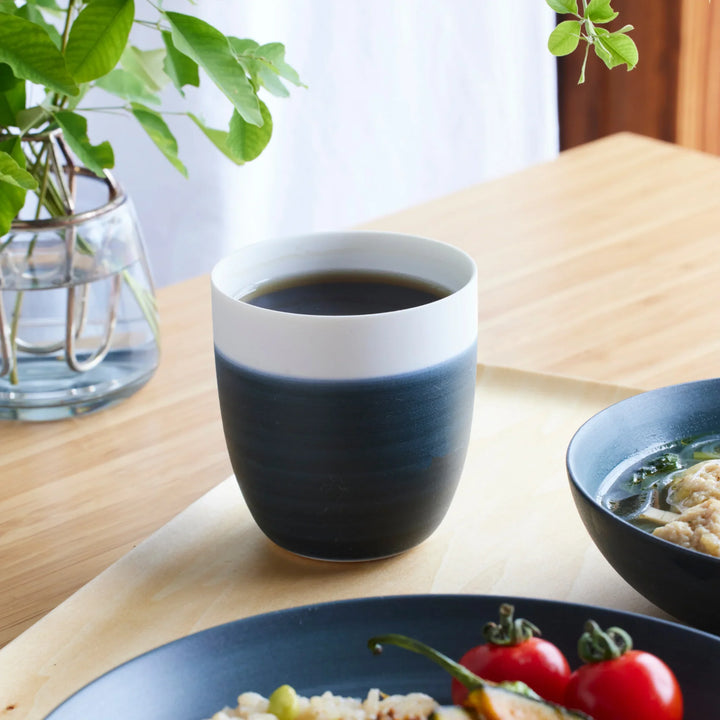 Japandi tea cup in navy and white, placed on a wooden table beside a meal and fresh greens.