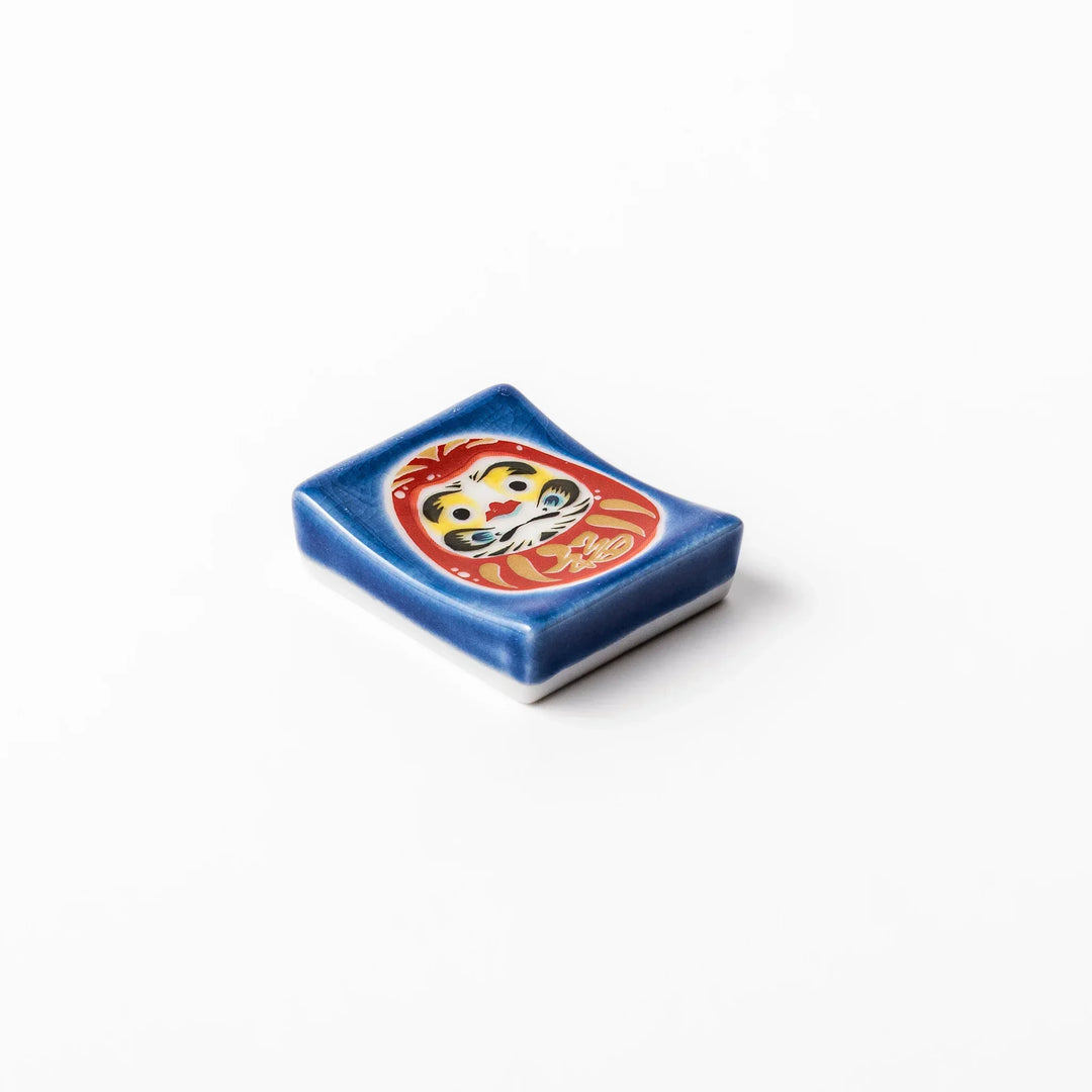 Rectangular Japanese ceramic chopstick rest with red Daruma doll on blue background symbolizing good luck.