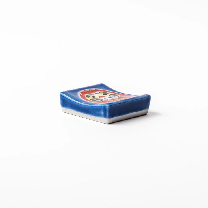 Rectangular Japanese ceramic chopstick rest with red Daruma doll on blue background symbolizing good luck.