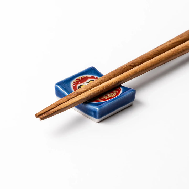 Rectangular Japanese ceramic chopstick rest with red Daruma doll on blue background symbolizing good luck.