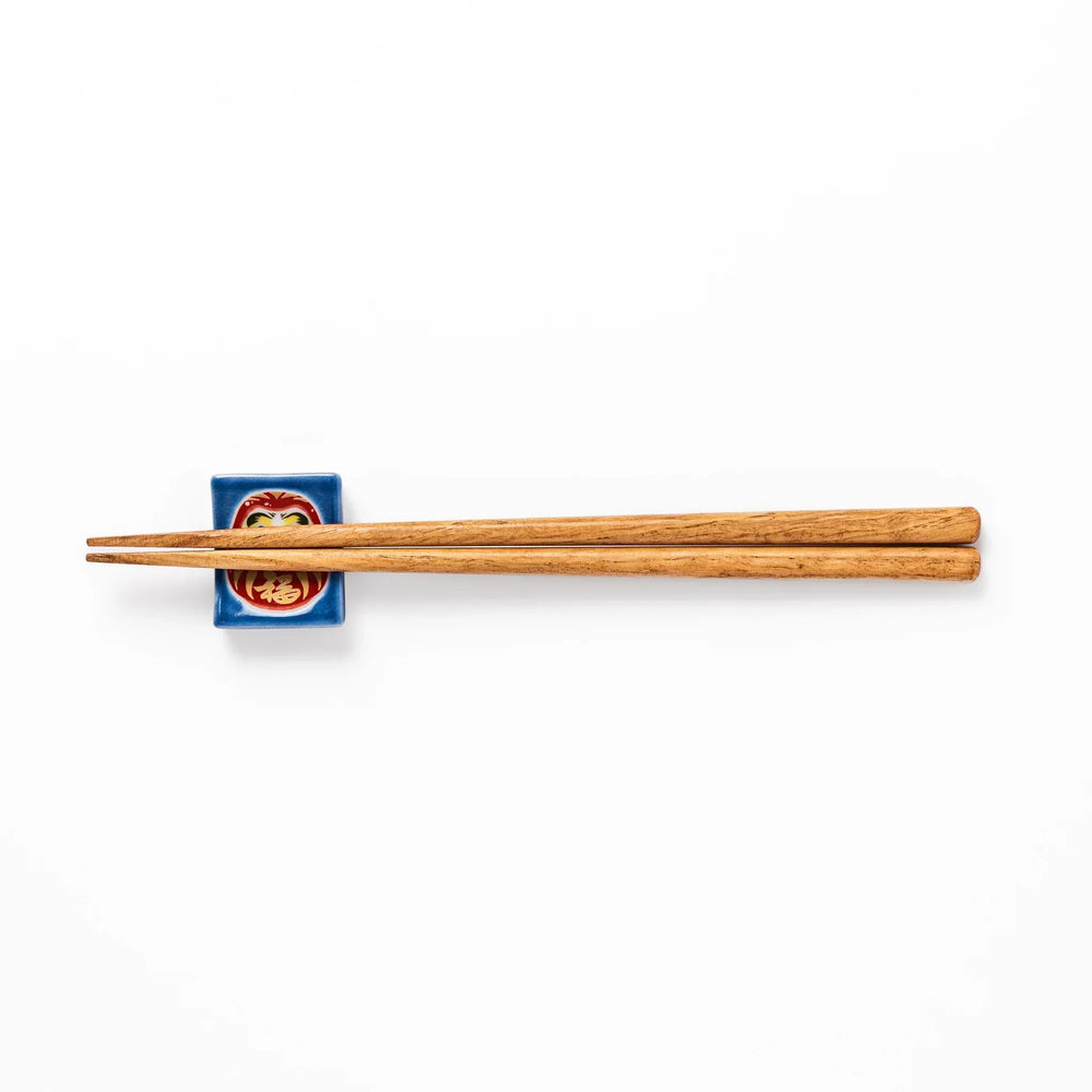 Rectangular Japanese ceramic chopstick rest with red Daruma doll on blue background symbolizing good luck.