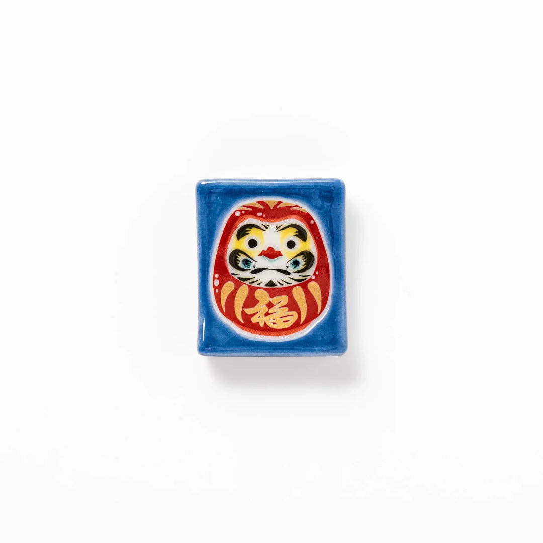 Rectangular Japanese ceramic chopstick rest with red Daruma doll on blue background symbolizing good luck.