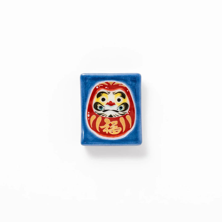 Rectangular Japanese ceramic chopstick rest with red Daruma doll on blue background symbolizing good luck.