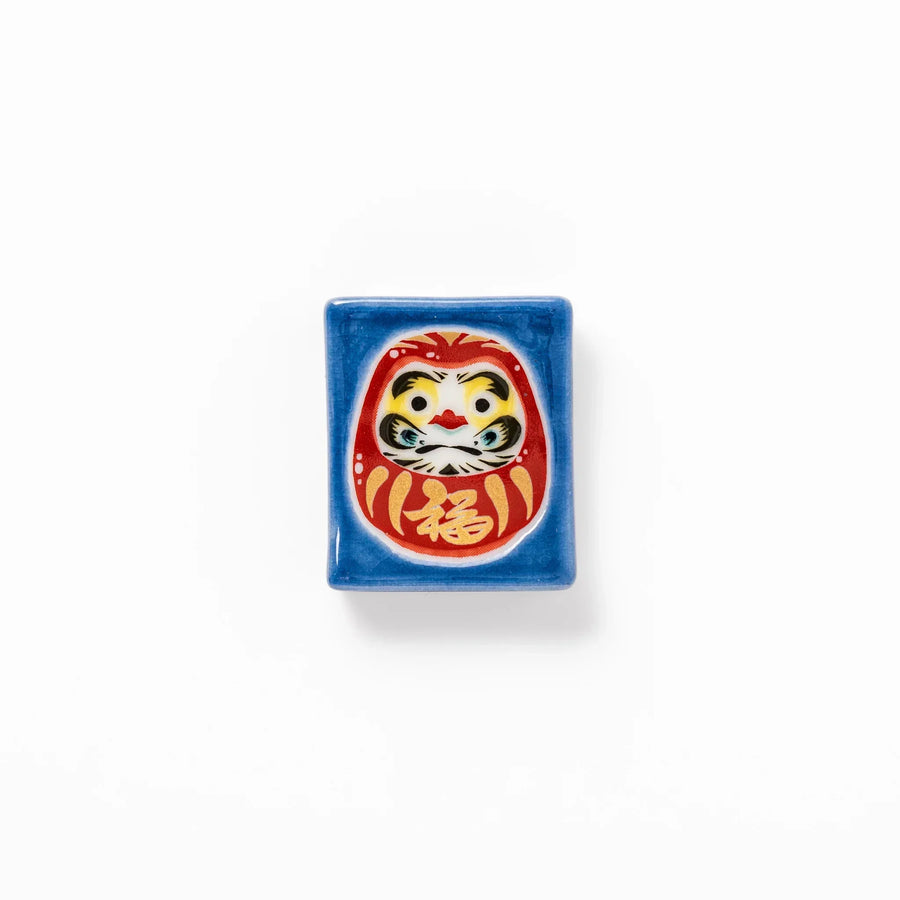 Rectangular Japanese ceramic chopstick rest with red Daruma doll on blue background symbolizing good luck.
